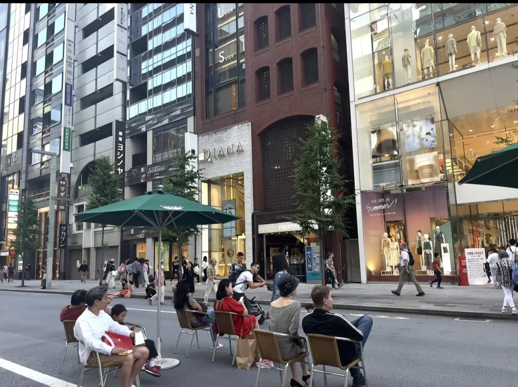 Ginza open street people sitting on chairs facing luxury department stores. A green umbrella standing on the street in Ginza.